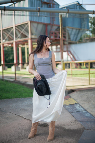 Woman holding a black cowgirl hat and a white skirt in an outdoor setting with industrial background